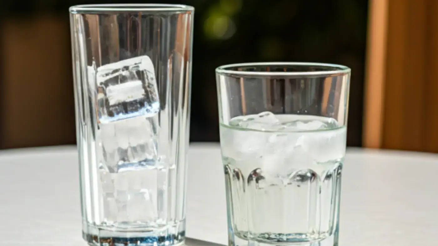 Two tall glasses filled with ice cubes are placed on a white table. The glass on the left has two crescent-shaped ice cubes inside. The glass on the right is filled with water and has several crescent-shaped ice cubes floating in it. The table is outdoors in a sunny location, with the shadows of the glasses cast on the table.