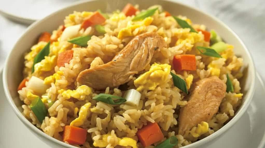 Steaming bowl of healthy chicken stir-fry over rice with scrambled egg, diced carrots, and fresh green onions.