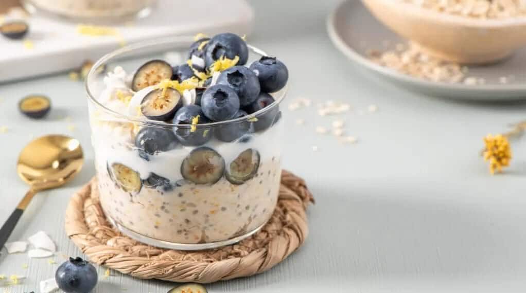 High protein meals with layered overnight oats, chia seeds, yogurt, sliced figs, fresh blueberries, and lemon zest in a clear glass jar on a woven coaster.