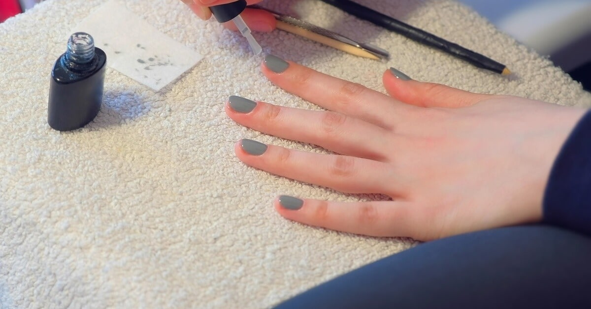 A close-up of someone painting their nails at home with grey gel nail polish. They're putting a top coat on.