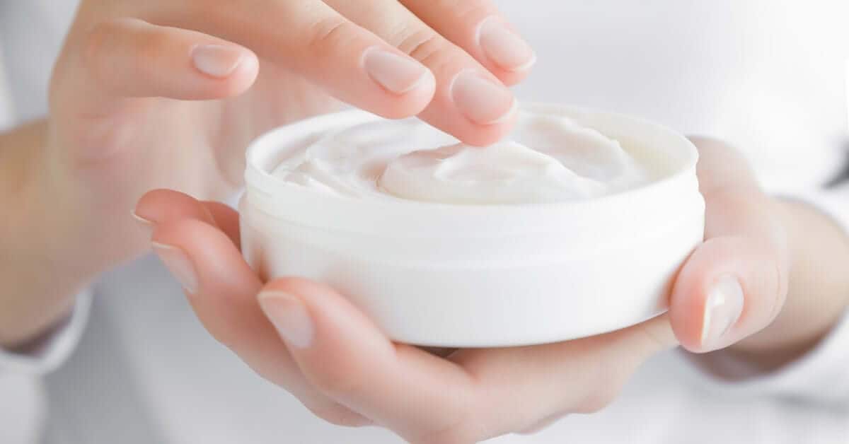 A person in a white long-sleeve shirt applies moisturizing cream from a small white round jar to their hands.