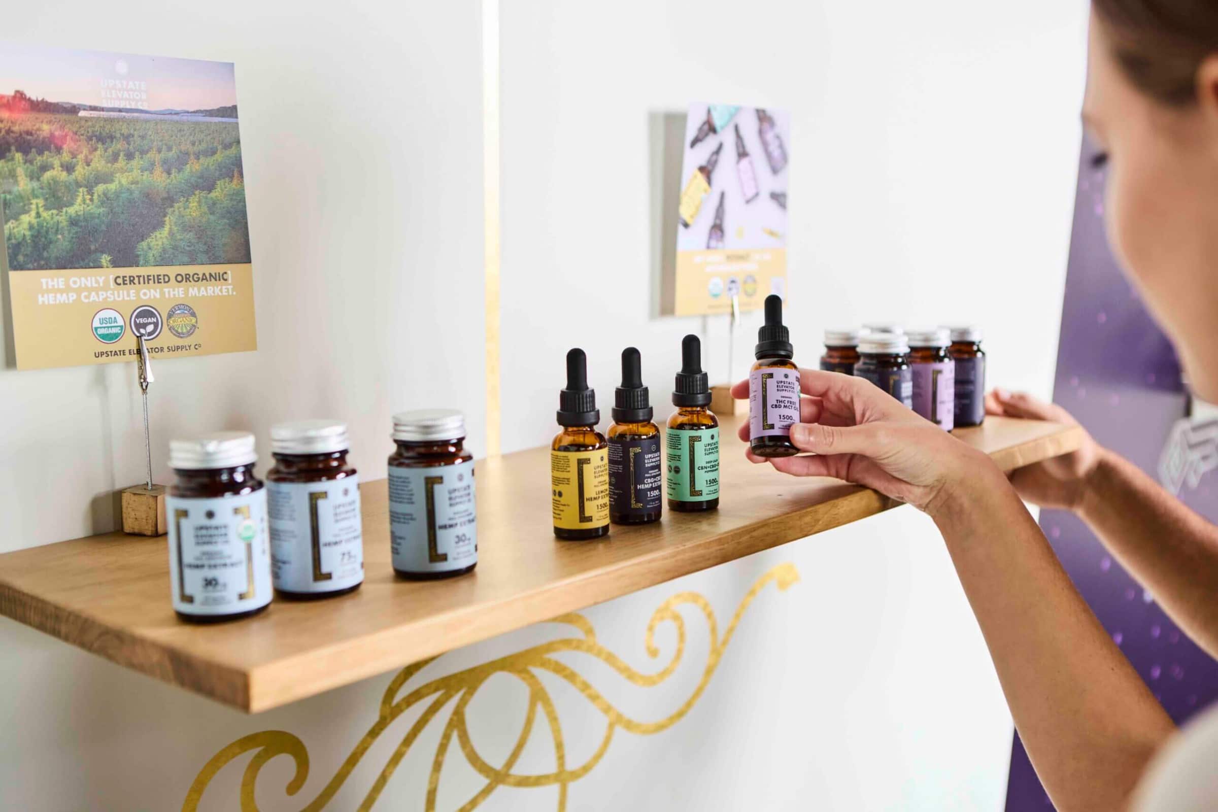 Hand picking up a dropper bottle from a wooden wall shelf of tinctures and capsule jars