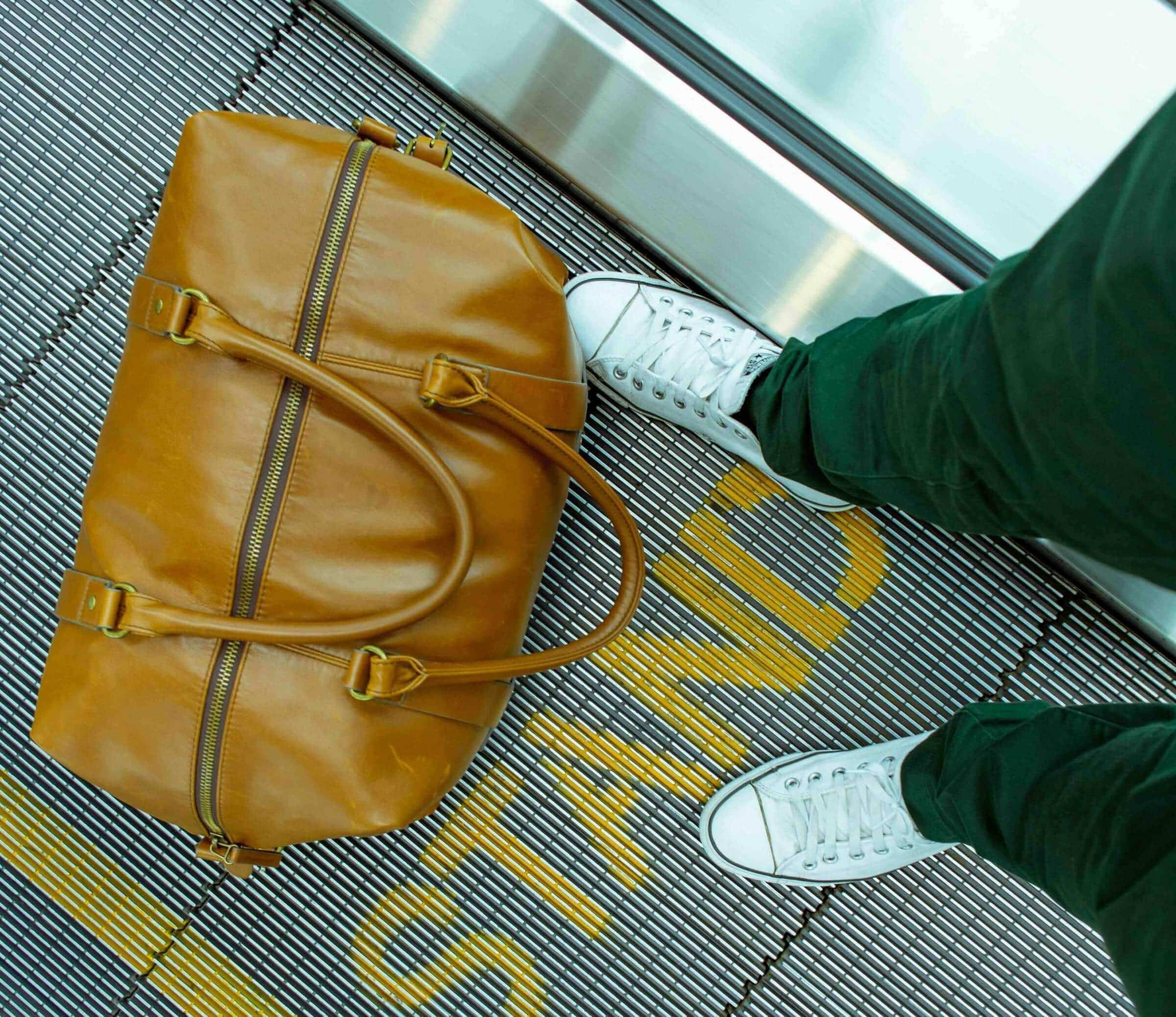 Traveler in white sneakers on an airport moving walkway beside a tan leather duffel bag