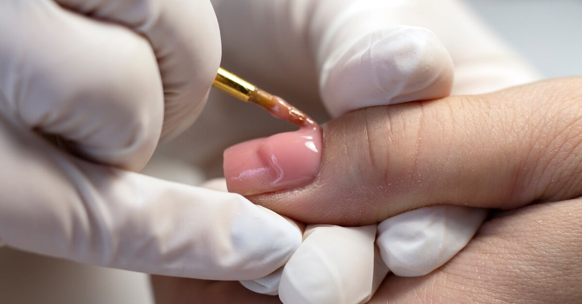 A gloved nail technician building up the surface of a client's nail with a thick, light pink gel topper that strengthens the nail.