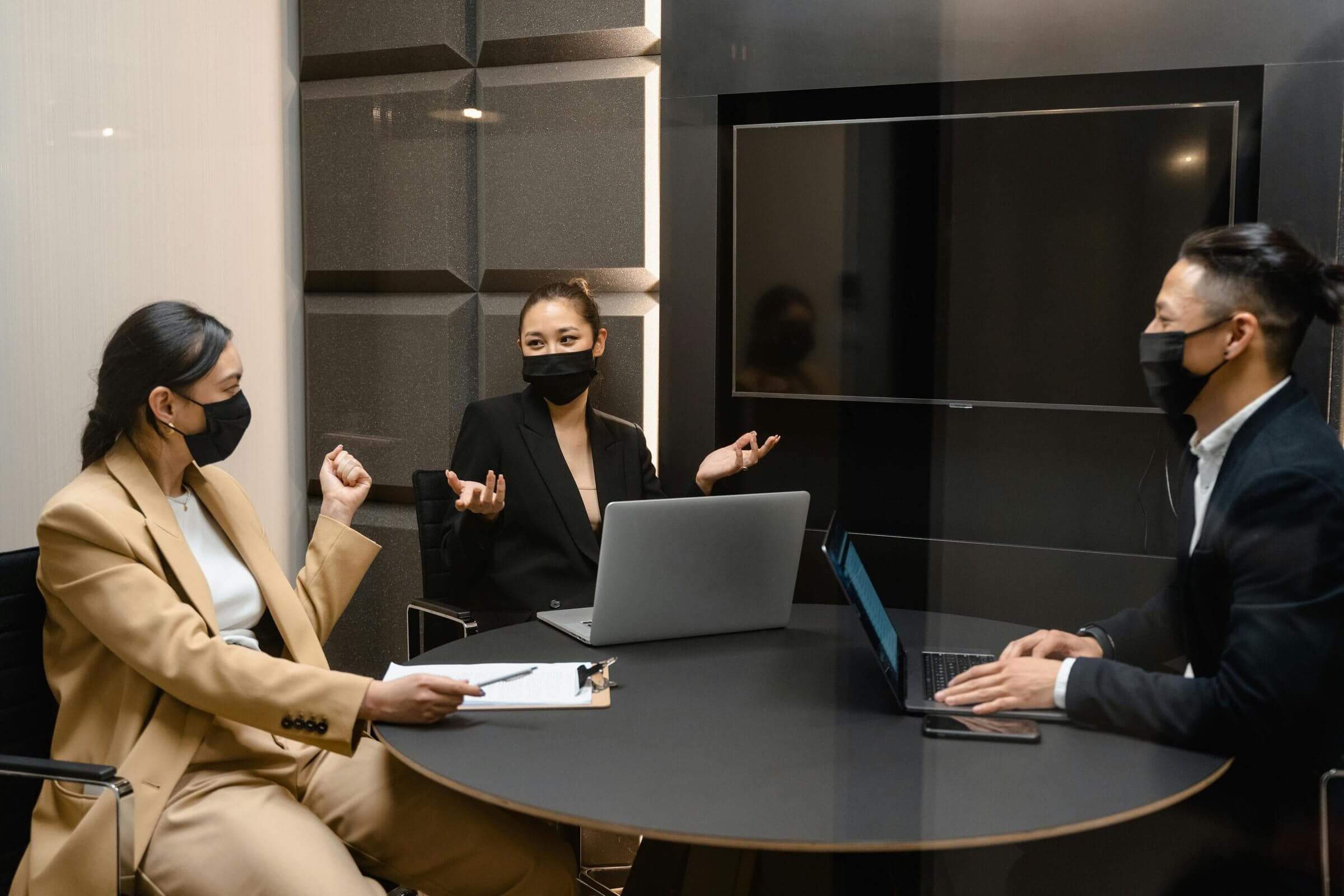 Three business professionals wear face masks during a meeting, collaborating around a round table indoors.