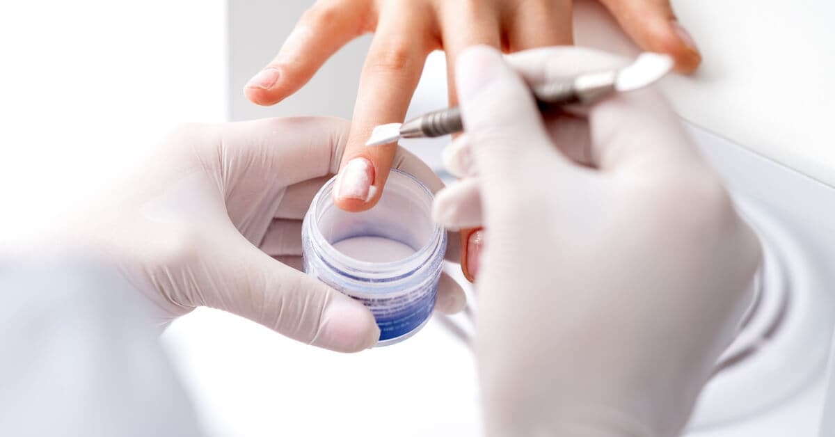 A close-up of a nail tech's gloves applying acrylic powder on the nails of a young person in a beauty salon.