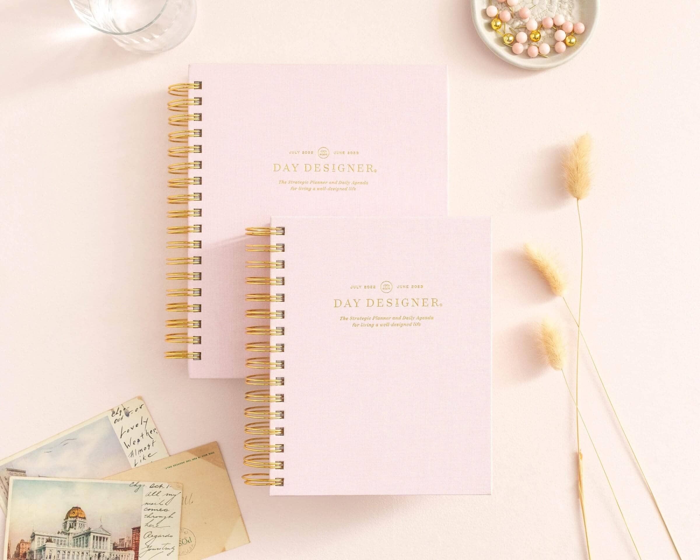 Which Planner Is Right For You?, stacked blush pink planners to show sizing options, dried wheat, dish with beads, postcards, water glass, beige background