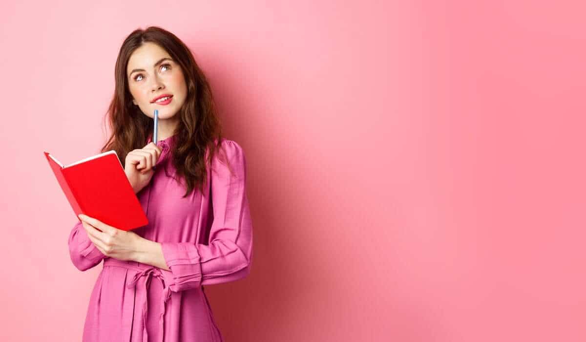 Thinking woman holding red notebook against pink background