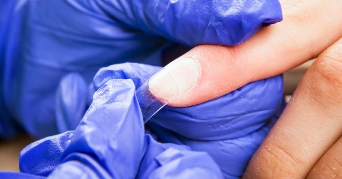 A nail technician gluing an acrylic nail onto the prepped natural nail of a client while wearing blue gloves.