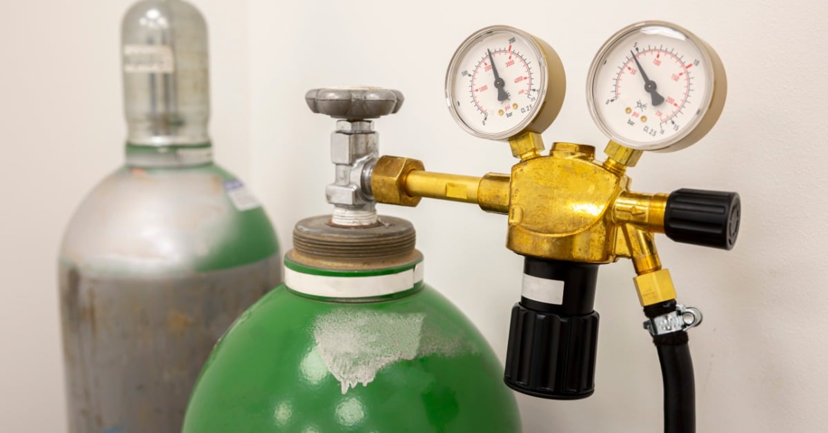 Green compressed gas cylinder with a brass pressure regulator, dual gauges, black hose, and a second cylinder behind it.