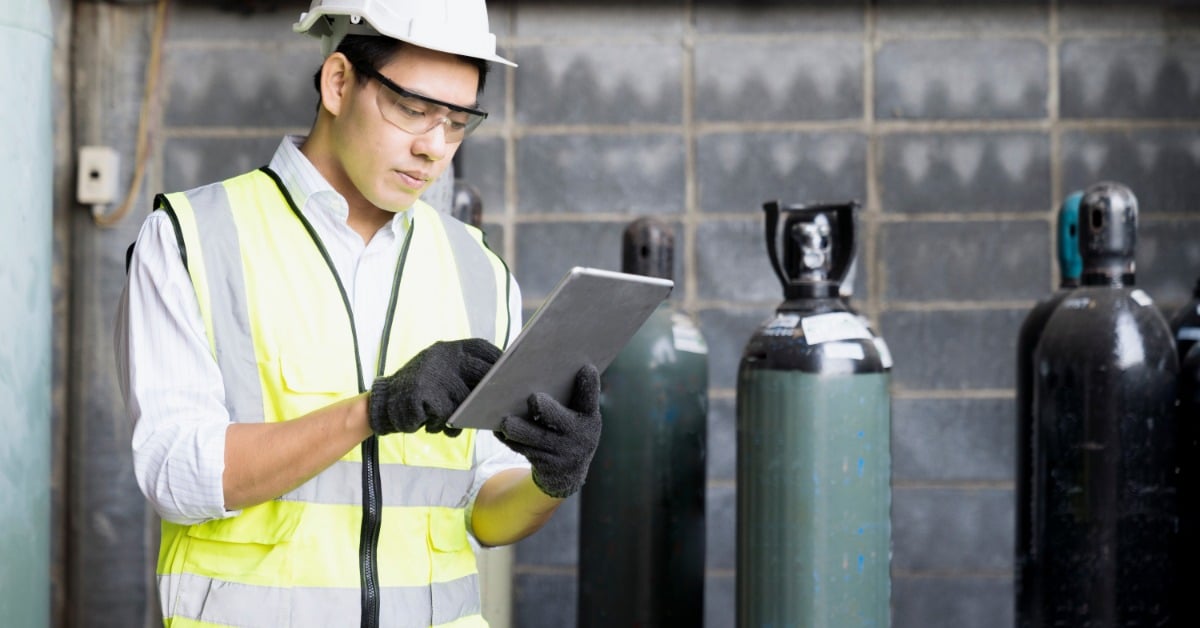 Worker in a hard hat and safety vest uses a tablet beside compressed gas cylinders in an industrial facility.