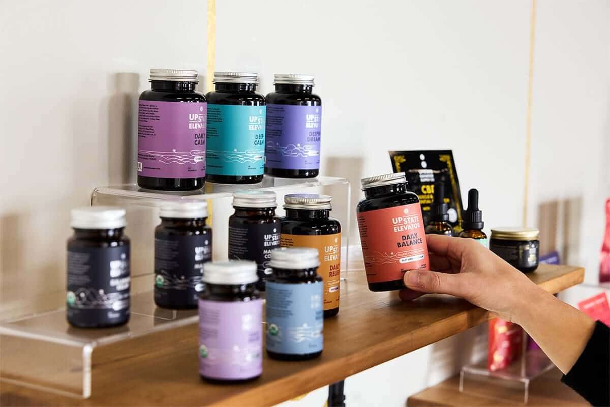 Hand lifting a jar of Daily Balance gummies from a shelf of Upstate Elevator jars and dropper bottles