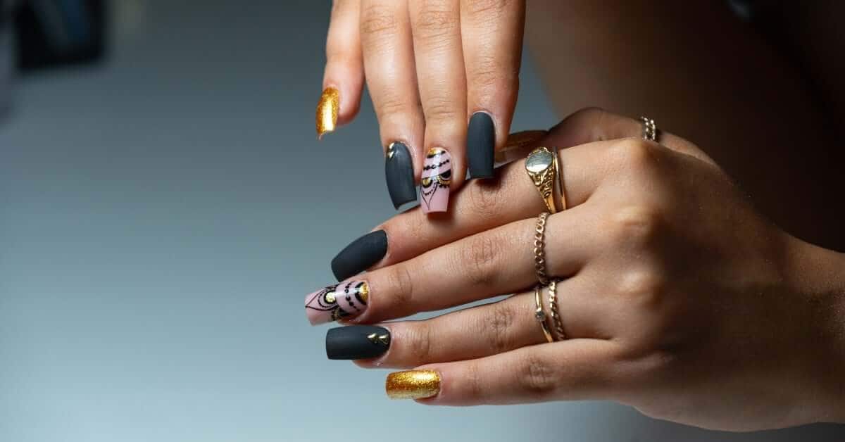 A pair of hands displays a stylish manicure with black, gold, and decorative nail art, adorned with gold rings and jewelry.