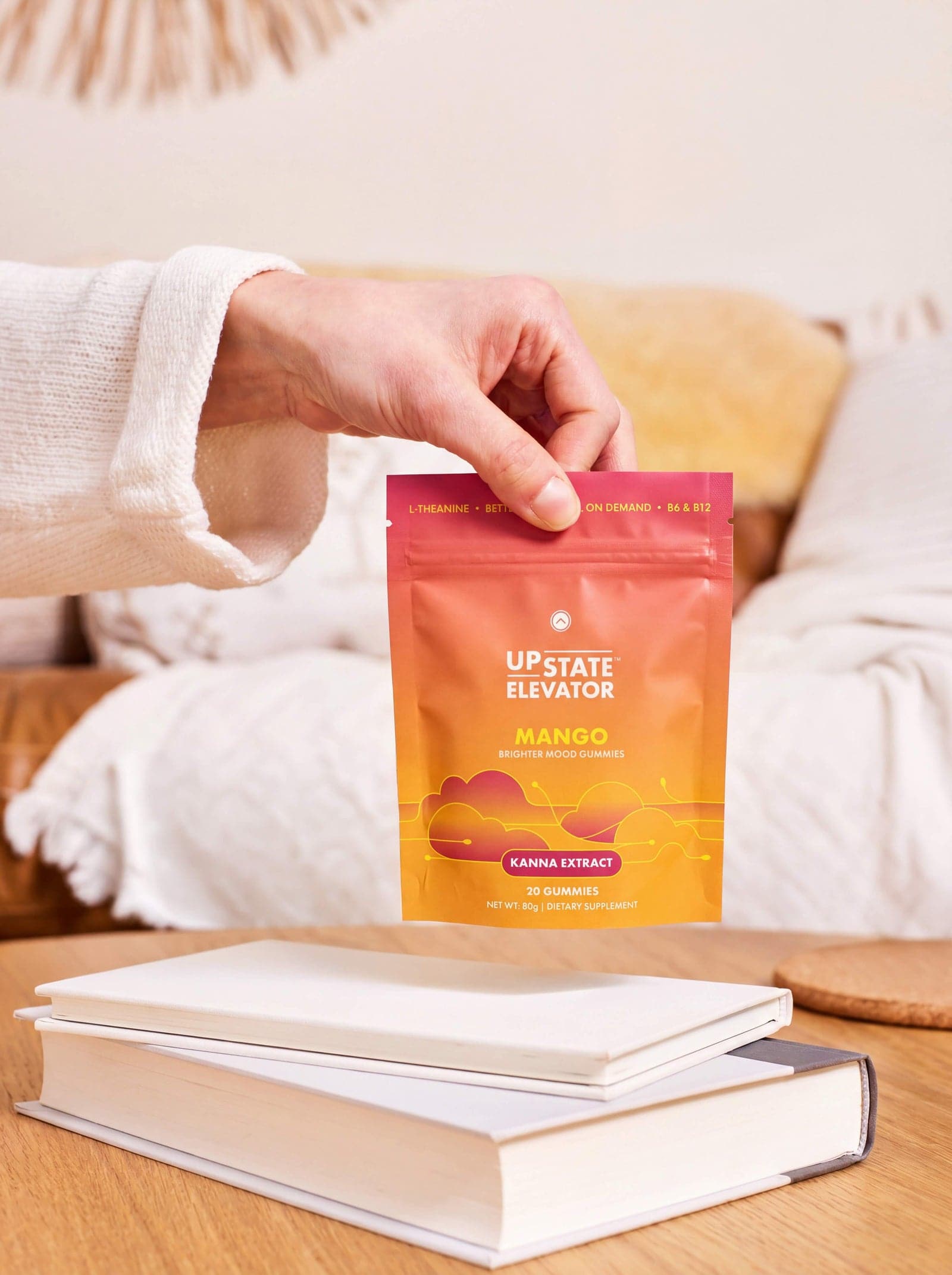 A hand lifting a pouch of Upstate Elevator Mango Brighter Mood Gummies above a stack of books, with a couch in the background.