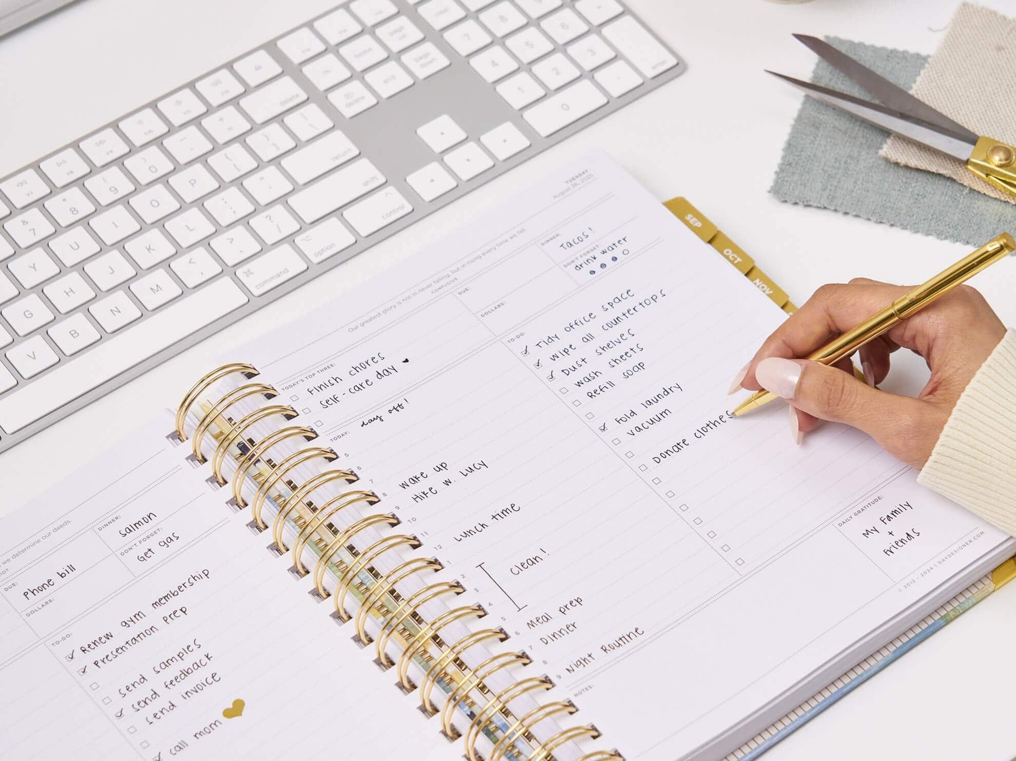 which planner format is right for you, hand with pen, writing on open book day designer planner, gold binding, keyboard, cloth, scissors