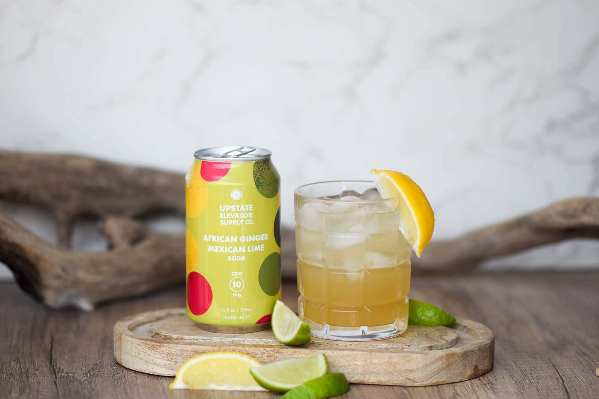Can of Upstate Elevator African Ginger Mexican Lime soda beside an iced cocktail with lemon on a wooden board