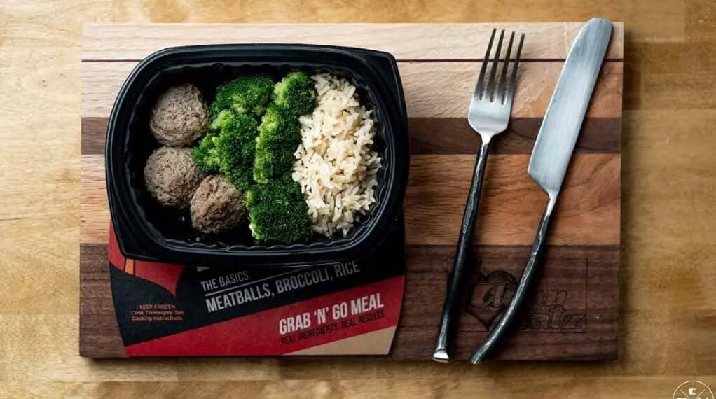 Portion control meal prep tray with meatballs, broccoli, and rice on wooden board with fork and knife.