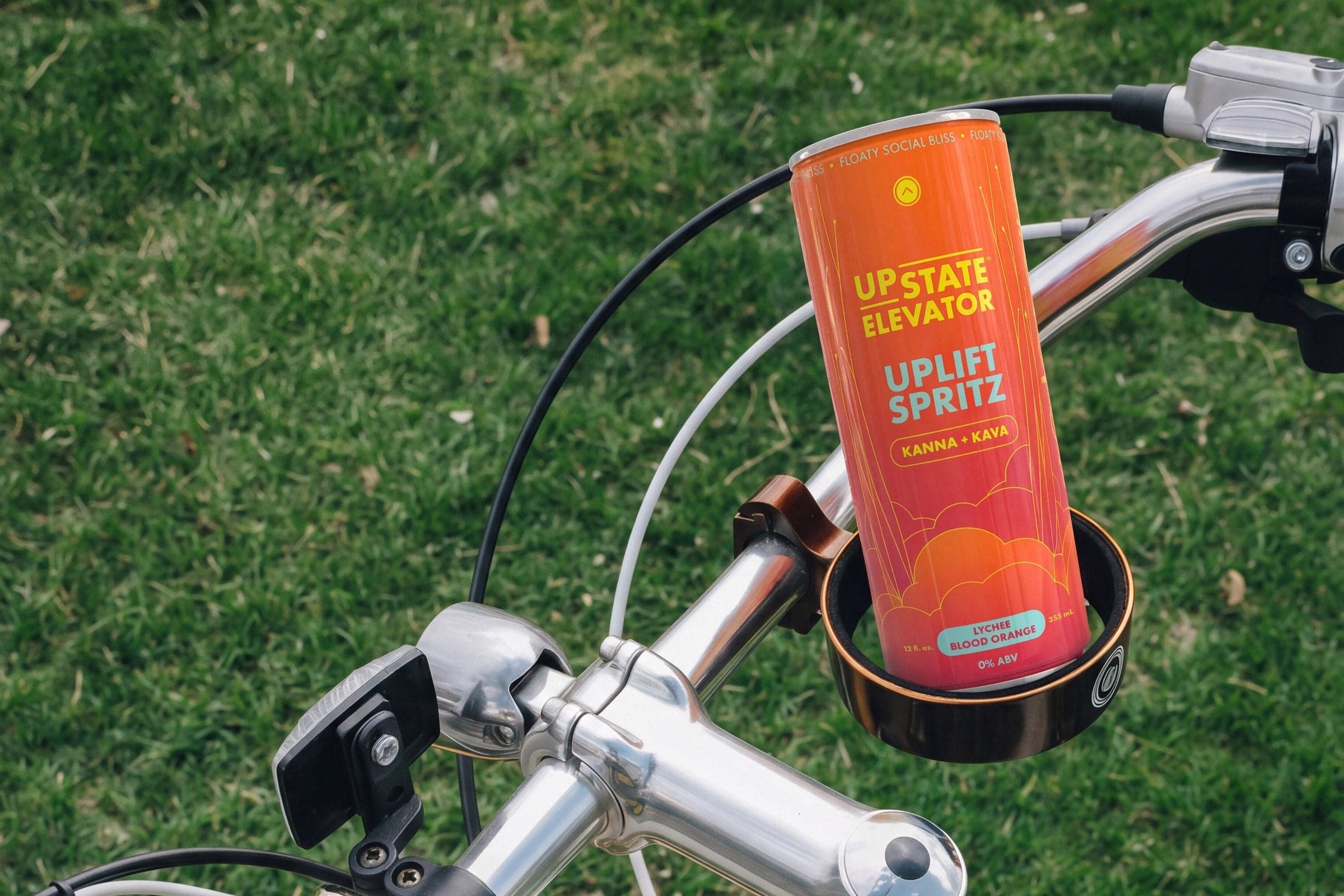 Can of Upstate Elevator Lychee Blood Orange Uplift Spritz in a cup holder on a bicycle handlebar above green grass