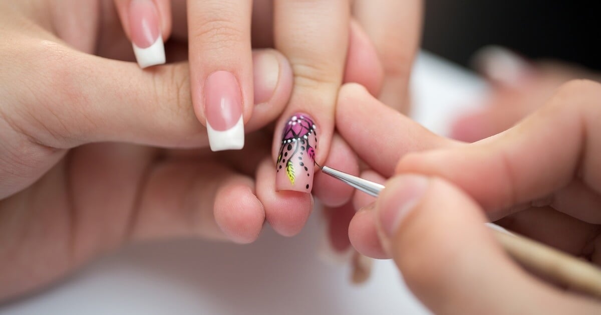 A person getting their nails done. Each nail has a classic French tip with someone painting a dream catcher on the ring finger.