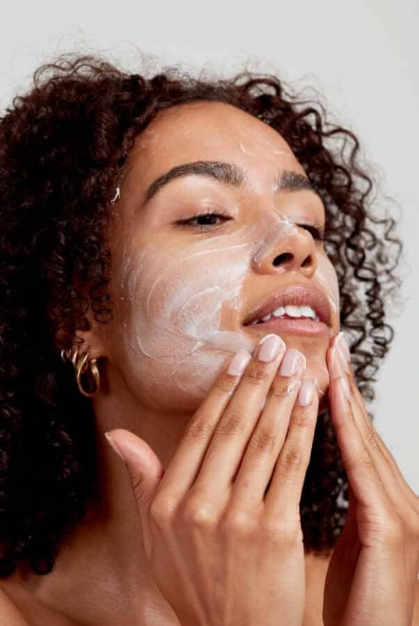 Woman with curly hair gently massaging white cream cleanser into her face with both hands