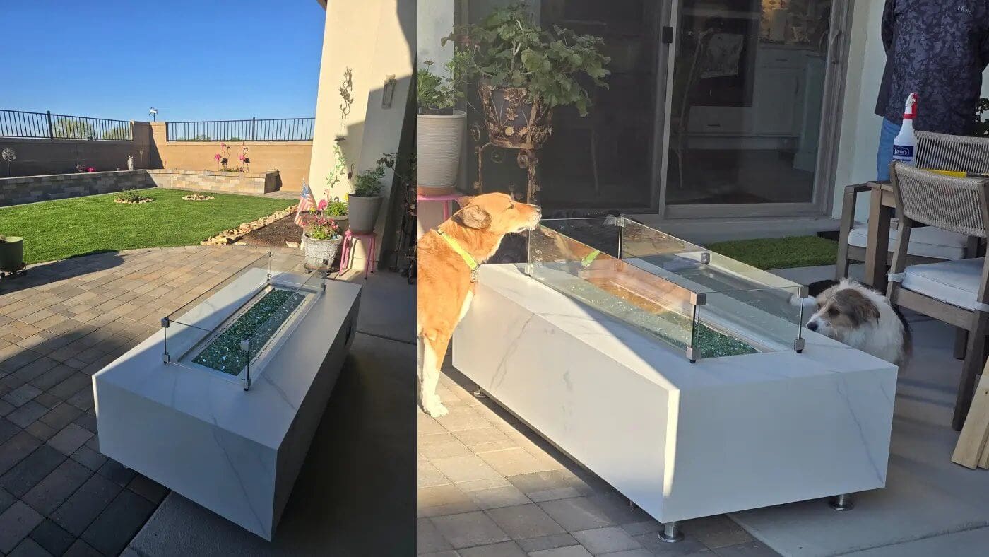 A long, white rectangular elementi fire pit table with a glass wind guard on a patio. Two dogs are nearby, and the background shows a sunny backyard with a lawn and fence.