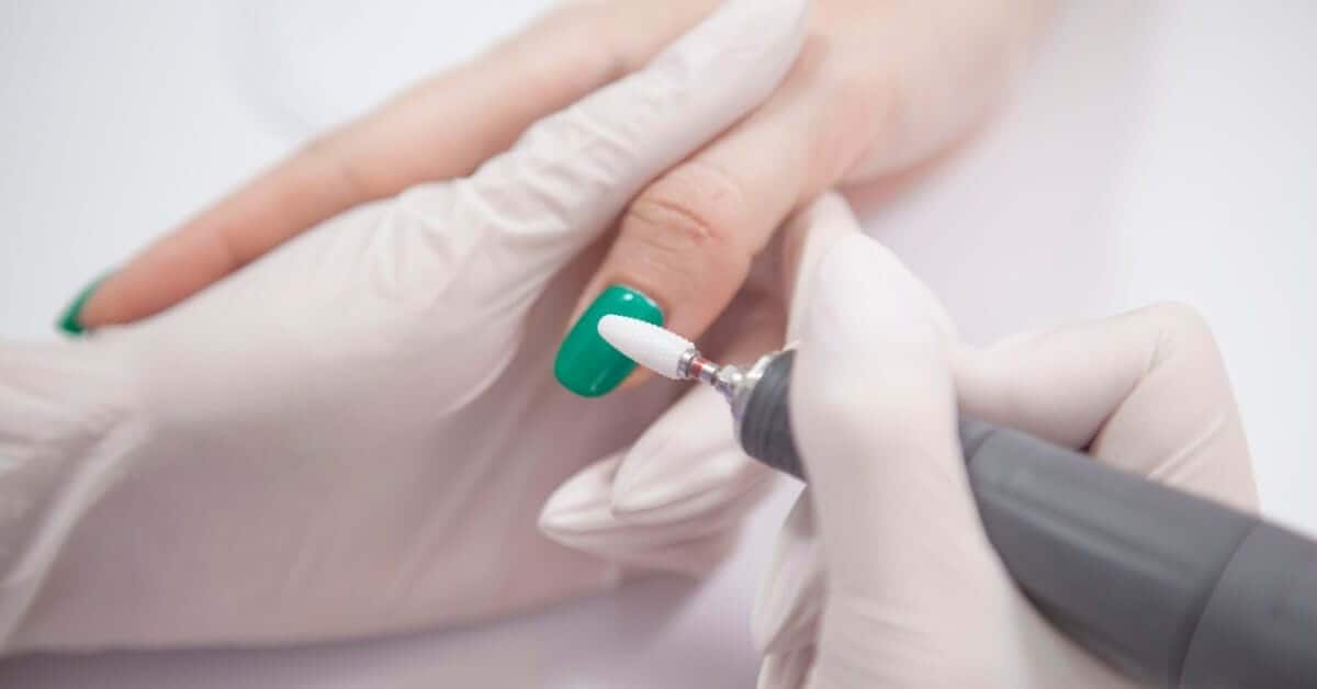 A nail technician in gloves preparing to use a tool to file off the cured green gel coating a client's thumb nail.