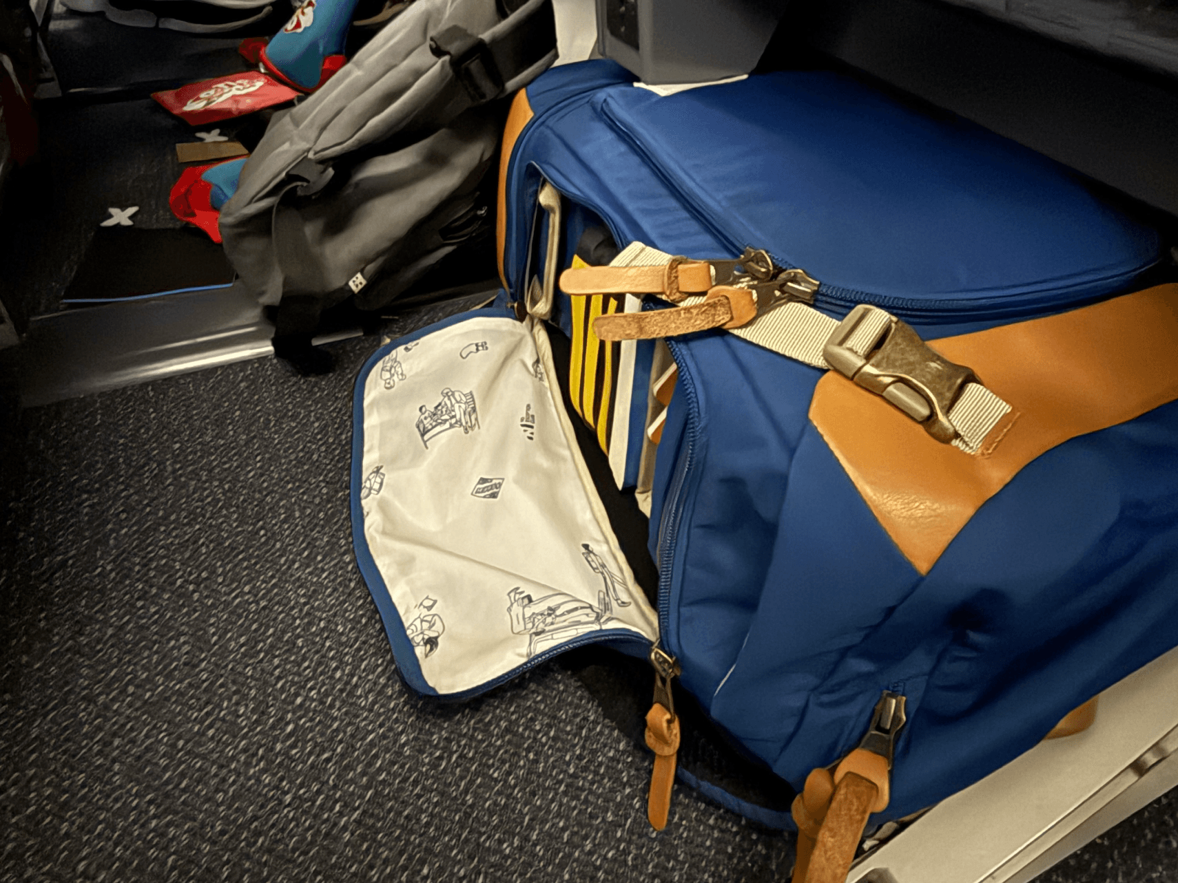 Blue Getaway diaper backpack lying sideways under an airplane seat, with its side panel unzipped to reveal books inside.
