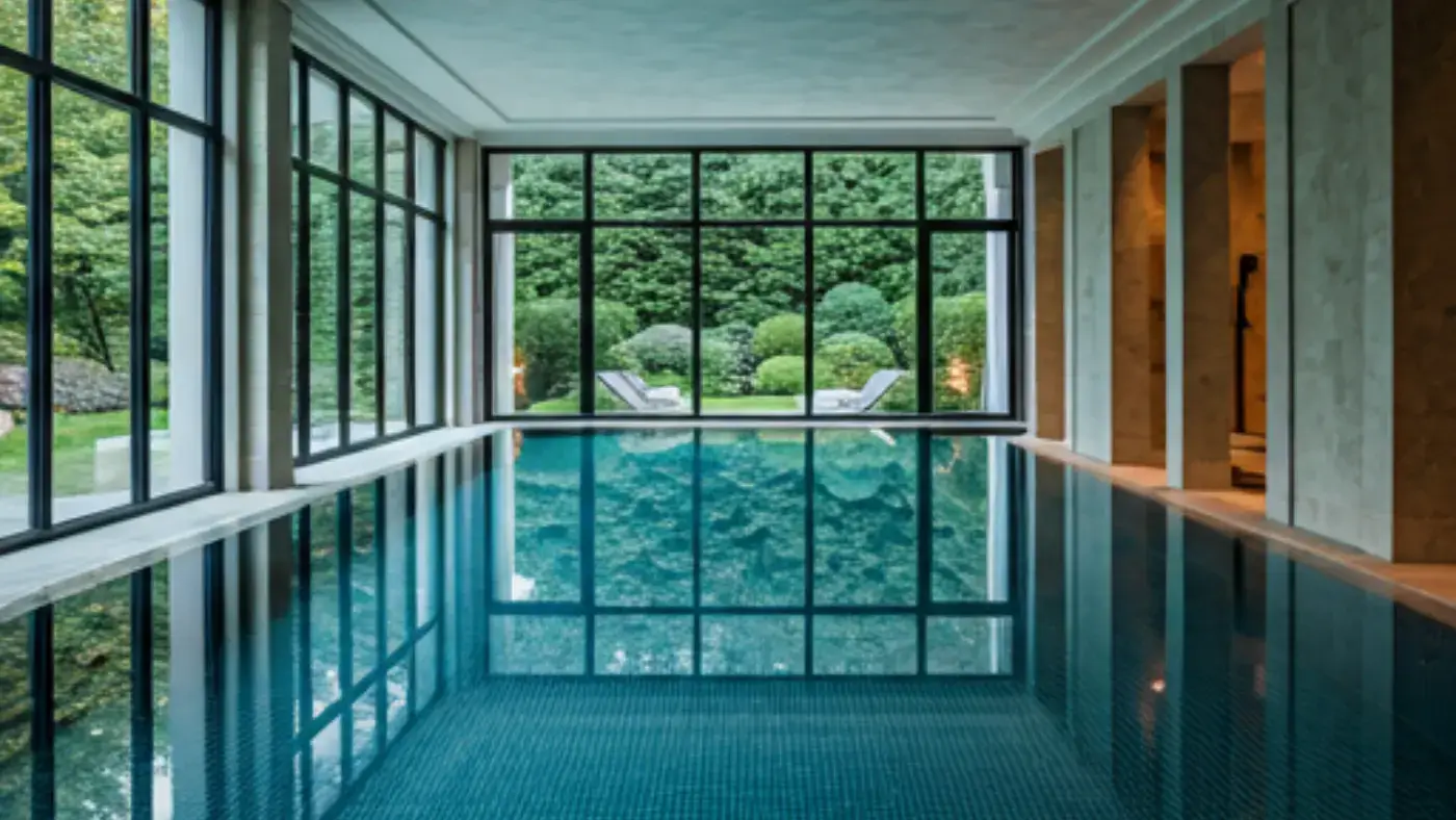 A tranquil indoor swimming pool, bathed in natural light from floor-to-ceiling windows, offering a serene view of a landscaped garden.