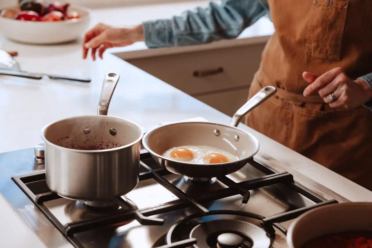 A person in a kitchen prepares eggs in a frying pan while a pot simmers nearby.