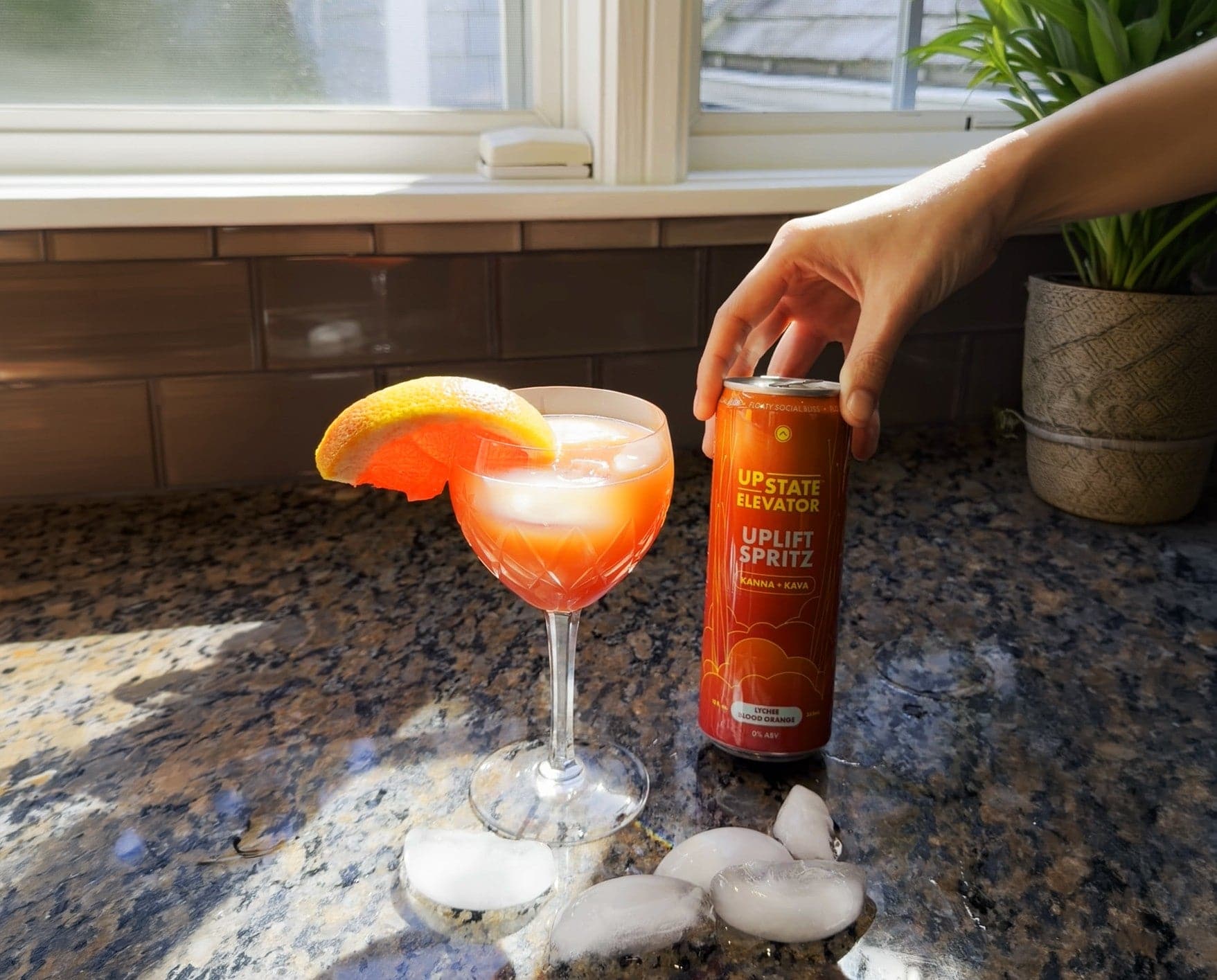 Pink Blood Orange Sunset mocktail in a cut-glass coupe with a blood orange wedge, beside a hand holding a can of Upstate Elevator Lychee Blood Orange Uplift Spritz on a sunny kitchen counter