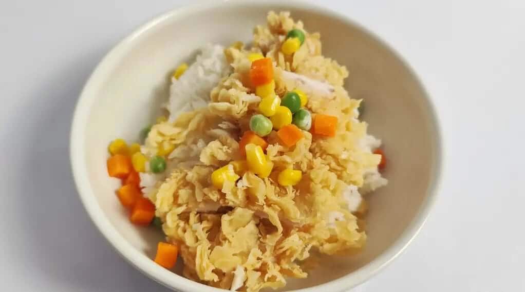 High protein chicken bowl with shredded egg-drop chicken over white rice topped with sweet corn, green peas, and diced carrots in a cream bowl.