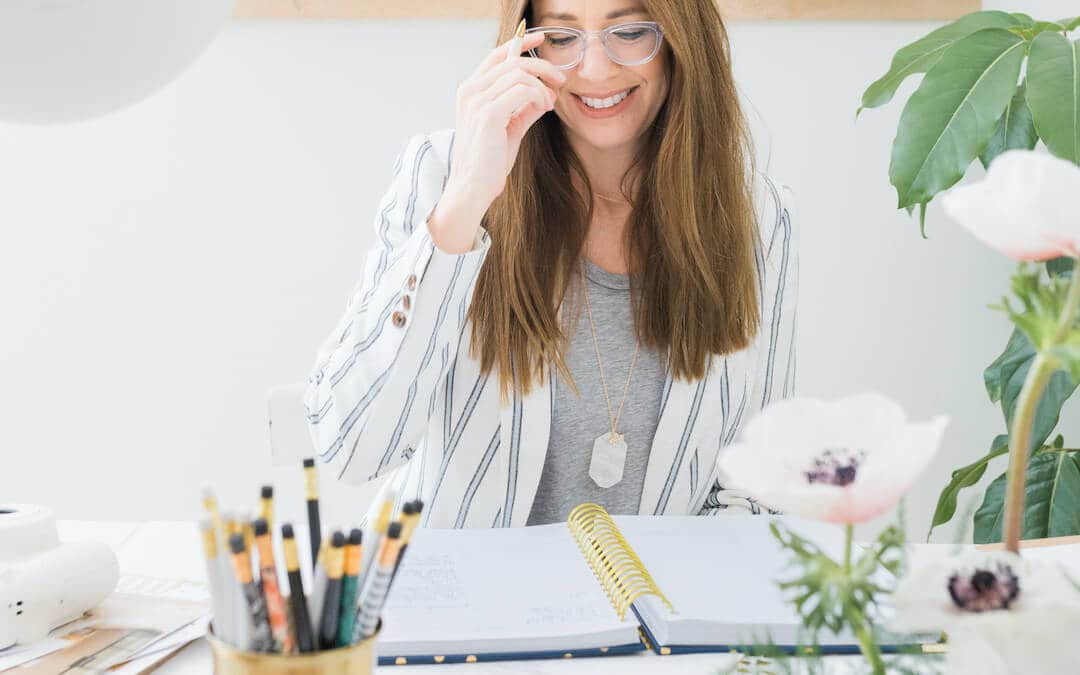 woman sitting at desk, holding eye glasses, open day designer planner, practicing efficiency flowers, plant, paint brushes in cup