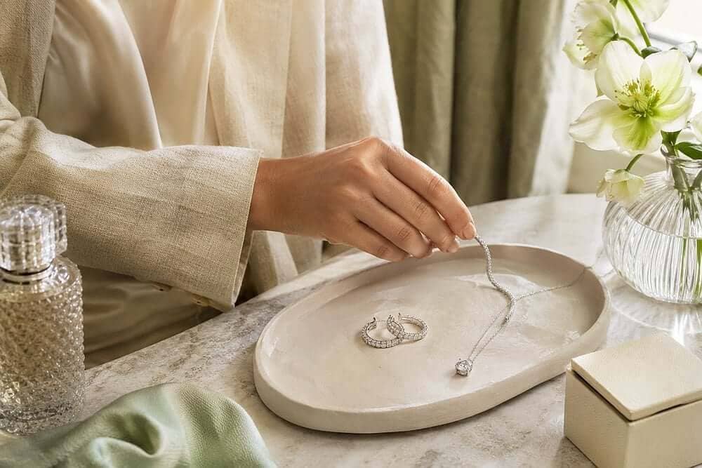 A hand selecting diamond jewellery from a dressing table with a cream blouse, pale linen sleeve, diamond pendant, hoops and bracelet in soft spring light.