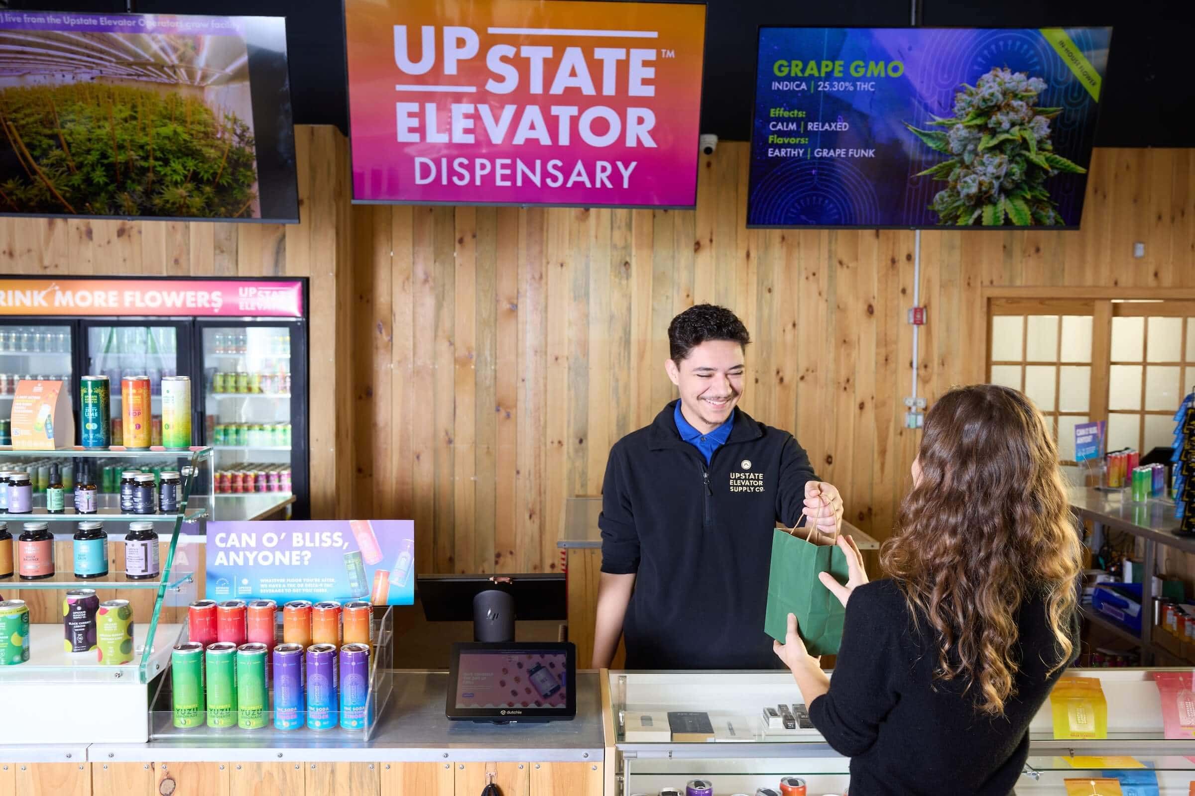 Smiling staff member handing a shopping bag across the counter to a customer at Upstate Elevator Dispensary