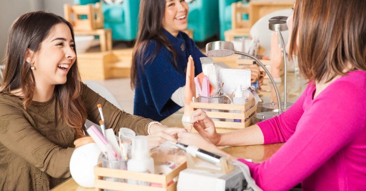 A group of women laughs and chats while enjoying nail treatments together in a vibrant salon setting.