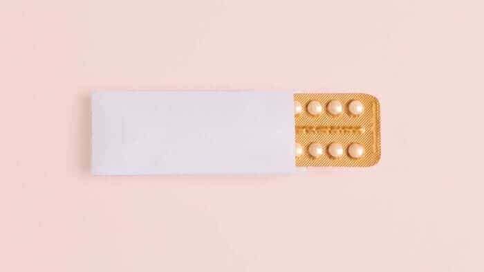 White pill packet with orange blister pack containing birth control pills on pink background