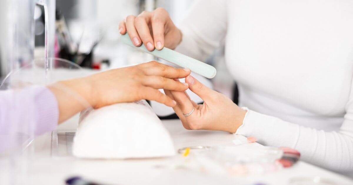 A manicurist shapes a young person's nails with a white nail file during a nail salon manicure session.