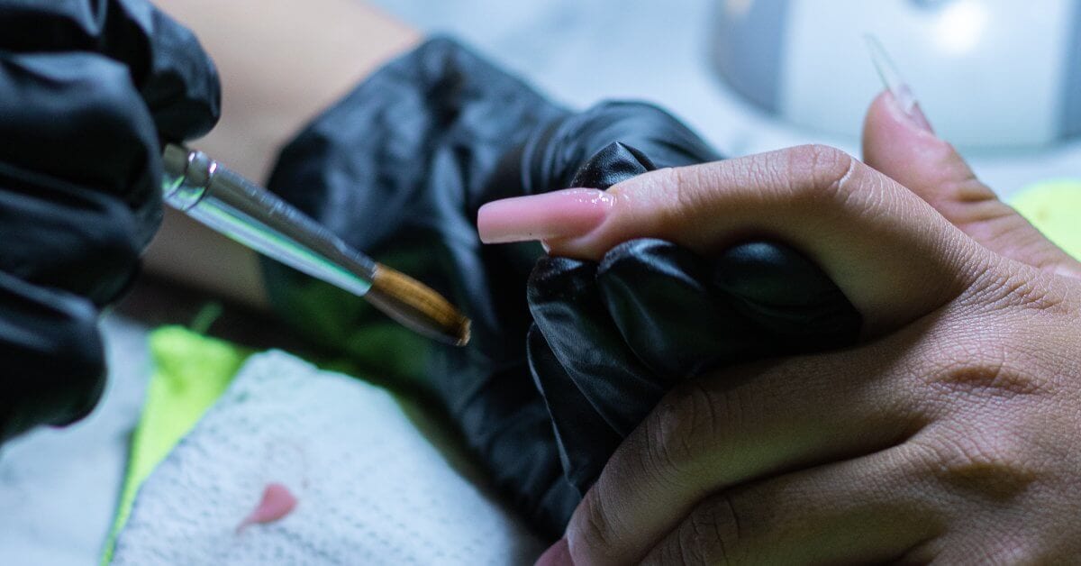 A close-up view of a black gloved hand holding the finger of another person; the other gloved hand holds a nail brush.