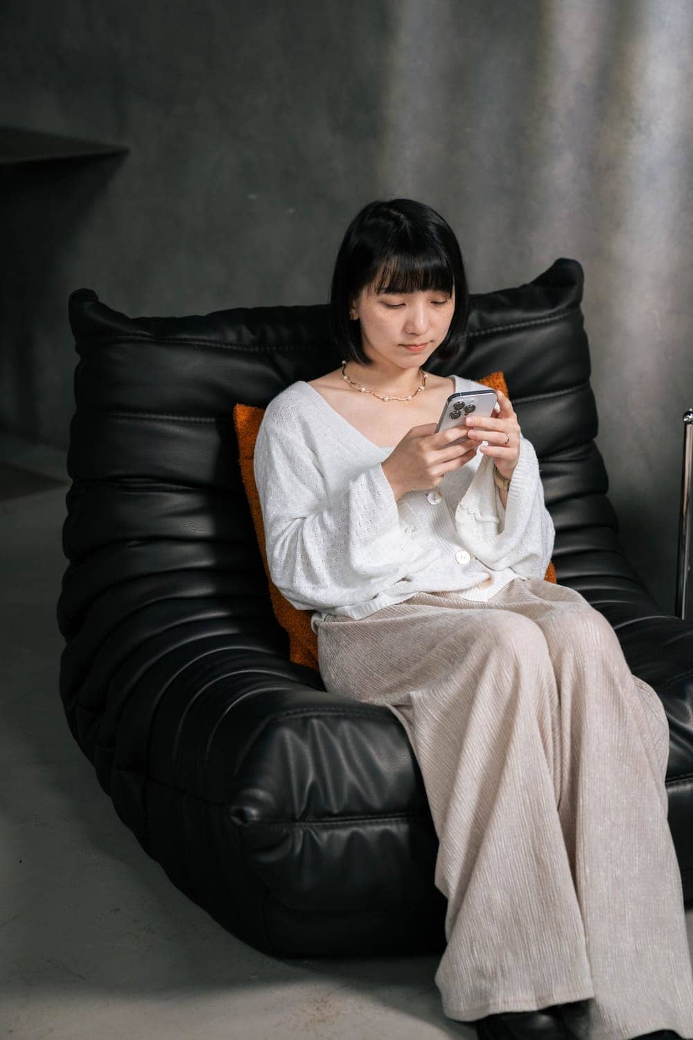 女性坐在黑色 Togo 單人沙發上使用手機，背靠橘色靠枕展現毛毛蟲沙發包覆放鬆的日常居家情境