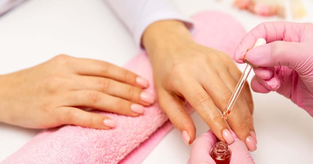 Close-up of a nail tech applying moisturizing fingernail oil on a person's hand resting on a pink towel.