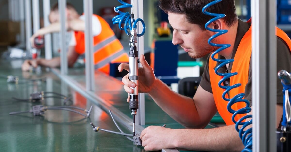 A man in an orange vest uses a handheld tool with a blue hose to assemble a component on a manufacturing line.
