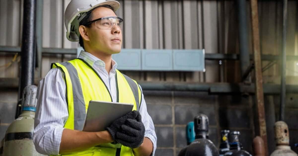 An industrial worker in a hard hat, safety vest, and gloves holds a tablet while standing in front of air receiver tanks.