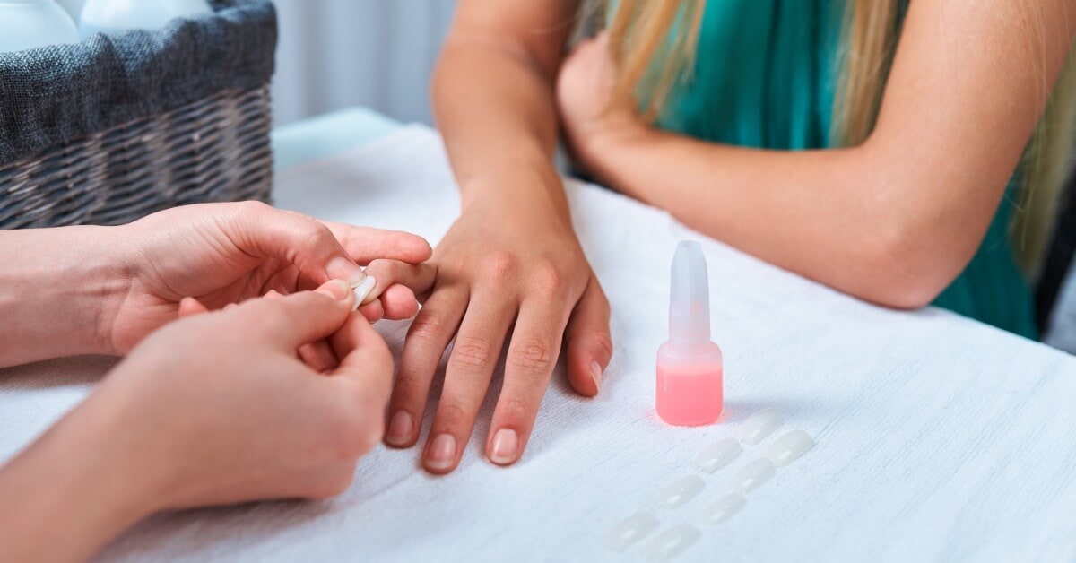 A client resting their hand on a white work surface while a technician applies a white plastic nail onto their natural nail.