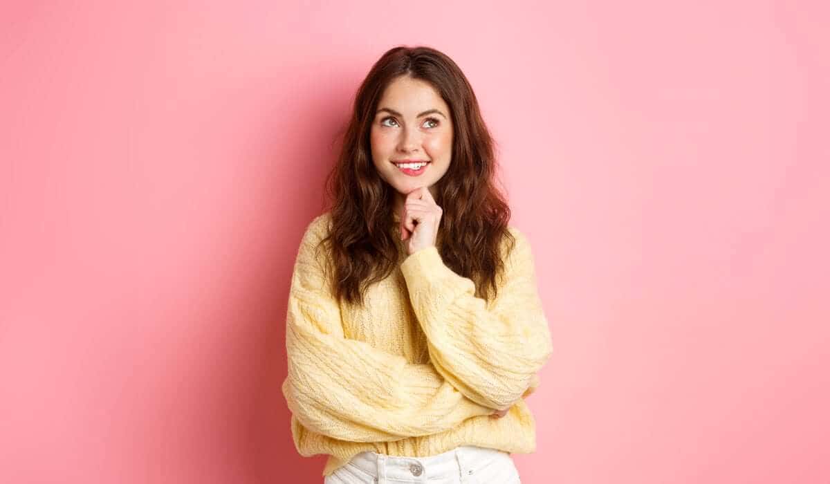Smiling woman in yellow sweater thinking against pink background