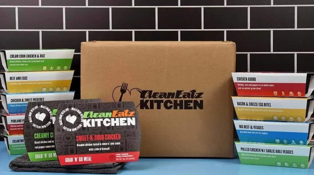 Clean Eatz Kitchen meal delivery box surrounded by individually packaged Grab 'N' Go meals like creamy chicken, sweet-and-sour chicken, and chicken kabob.
