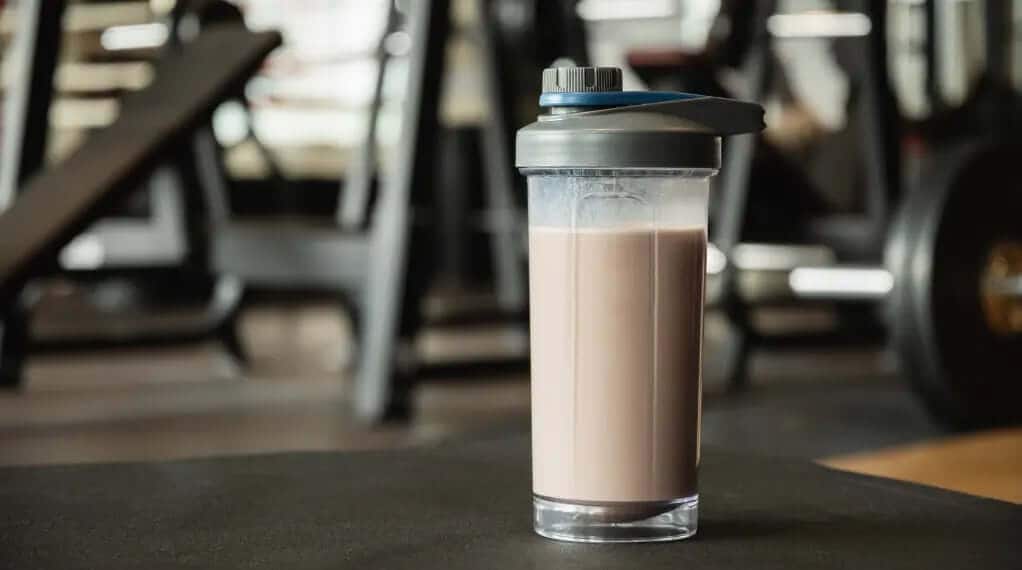 Protein shake in an open shaker bottle sitting on a gym floor surrounded by workout equipment.