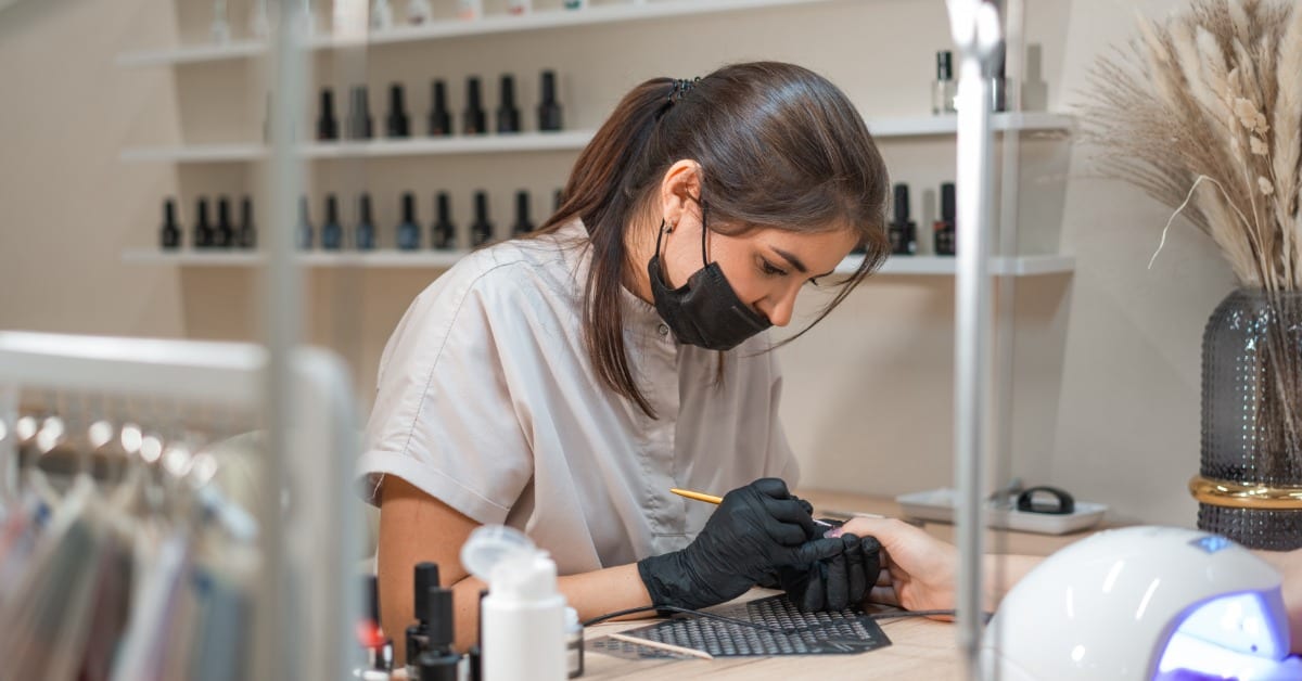 A female nail technician wears a black mask and black gloves while painting a client's fingernails.
