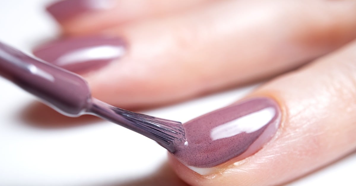  A close-up view shows a nail polish brush applying a purple polish to a fingernail. The hand rests on a white surface.