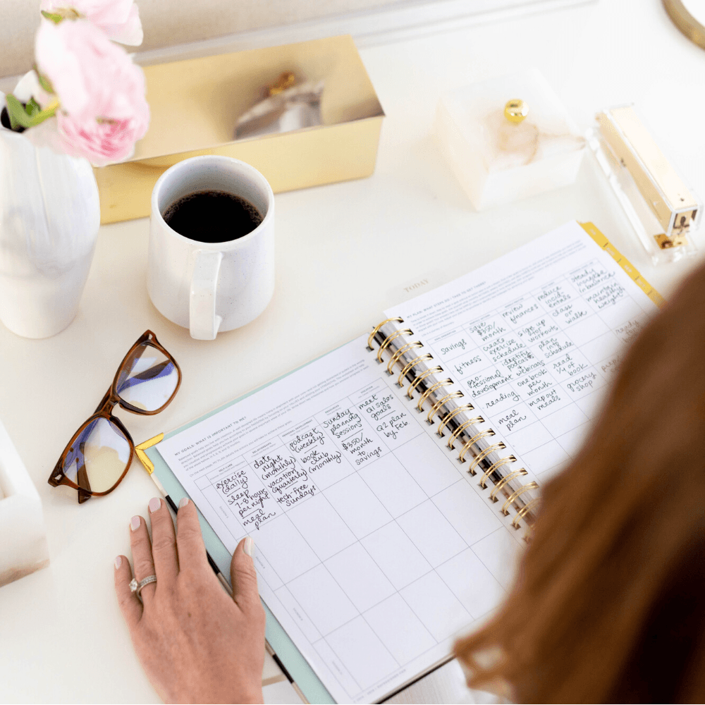 woman sitting at desk with black and white striped day designer planner, lamp, whiteboard with pictures, office accessories, achieve your goals,