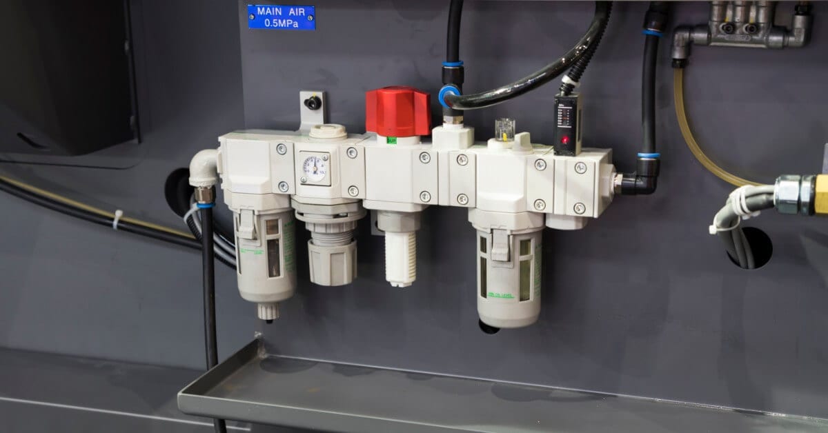A white FRL unit with several black tubes and a check valve is mounted to a gray piece of equipment.